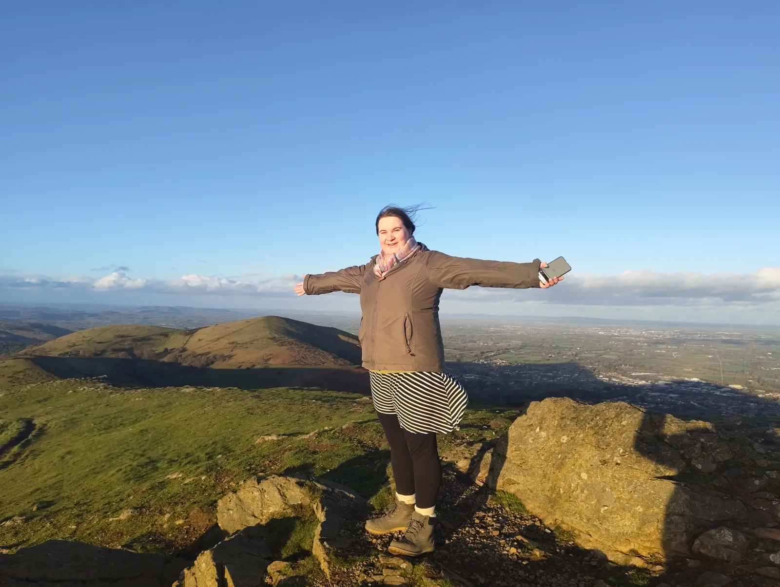 Alice standing on a sunny hilltop with her arms outstretched and the countryside behind her.
