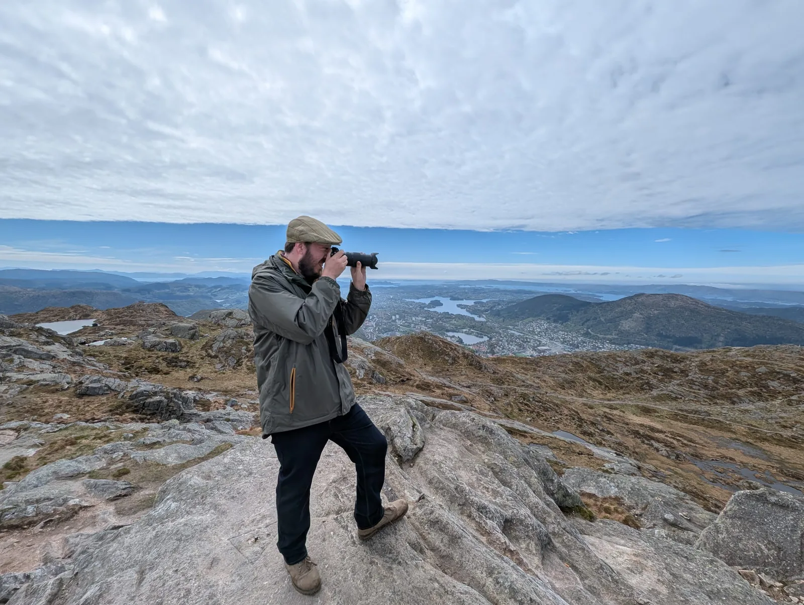 Robert taking a photograph from a rocky hilltop, with hills and water stretching out behind him.