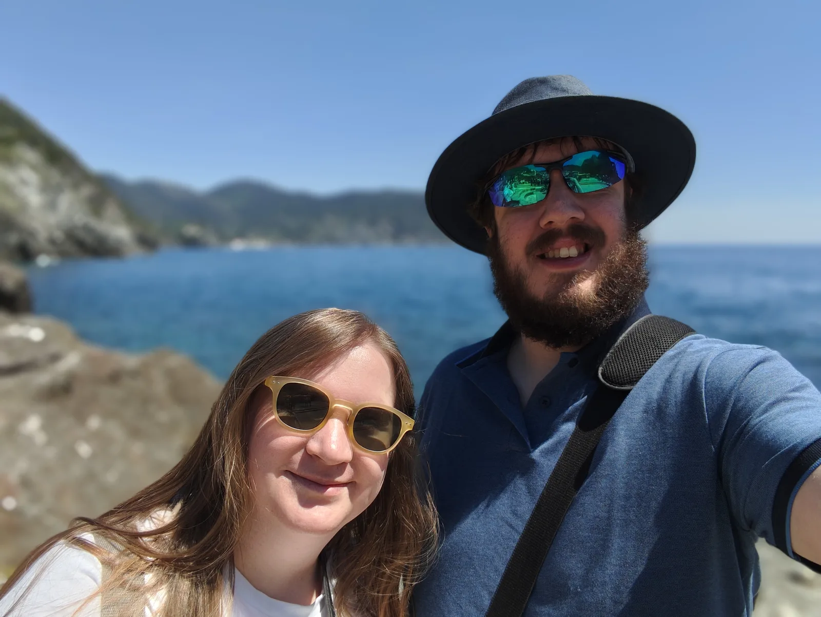 Alice and Robert wearing sunglasses beside the blue sea, with a rocky coastline behind them.