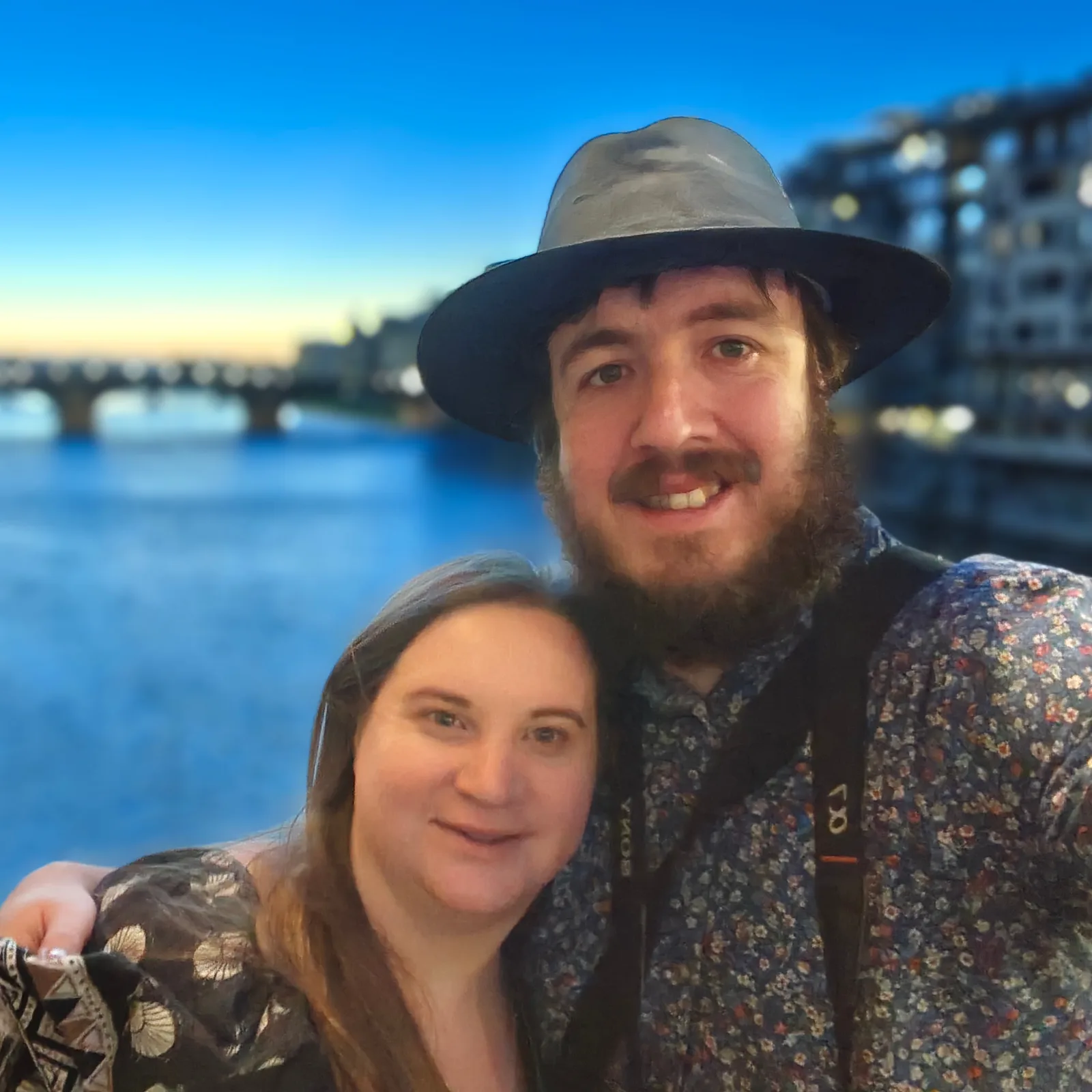 Robert and Alice smiling together beside a river, with a bridge behind them in the evening light.