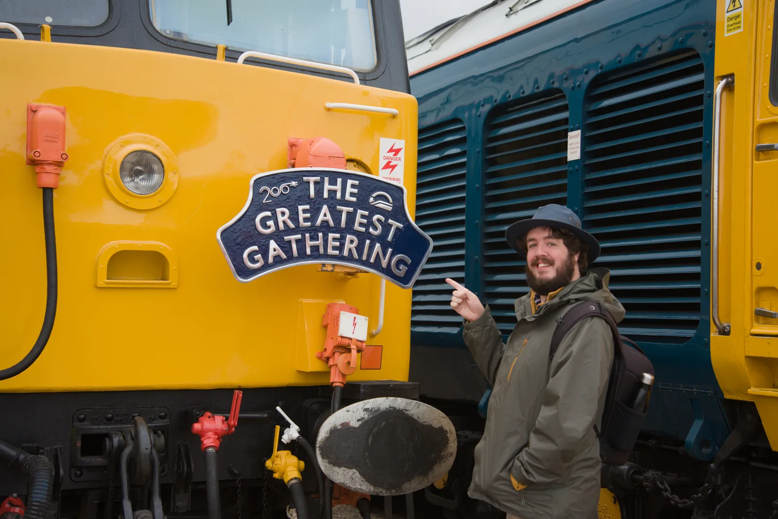 Robert smiling beside a yellow locomotive and pointing at The Greatest Gathering sign.